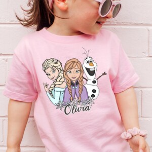 A young girl wearing a pink t-shirt with a graphic of the Disney characters Elsa, Olaf, and Oliva.