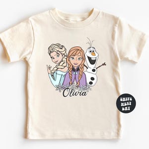 A white t-shirt with a graphic design featuring two cartoon characters, a girl and a snowman, along with the name "Olivia" written below them.