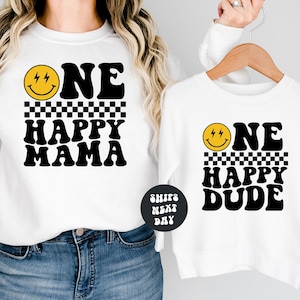 A woman wearing a white sweatshirt with a black and yellow smiley face design and the text "ONE HAPPY MAMA" printed on it, holding the sweatshirt up to the camera.