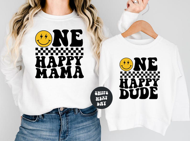 A woman wearing a white sweatshirt with a black and yellow smiley face design and the text "ONE HAPPY MAMA" printed on it, holding the sweatshirt up to the camera.