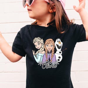 A young girl wearing a black t-shirt with a graphic of two cartoon characters, one of which is a snowman, and the other is a girl with blonde hair.