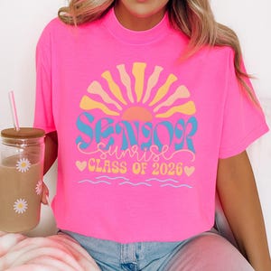 A woman wearing a pink t-shirt with a graphic design that says "SENIOR SUNRISE CLASS OF 2026".