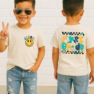 A young boy wearing a white t-shirt with a smiley face and the text "First Grade" printed on it, smiling and making a peace sign gesture.