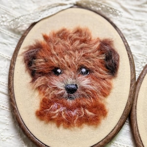 May include: An oval wooden embroidery hoop containing a needle-felted portrait of a fluffy, reddish-brown dog with dark ears, black eyes, and a black nose, set against a plain, light beige fabric background with a white lace ribbon at the top.