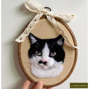 May include: An oval wooden embroidery hoop containing a needle-felted black and white cat portrait on beige fabric. The cat has a pink nose and white whiskers. A cream-colored lace ribbon is tied in a bow at the top. The text Le Cadeau Enchante is visible.