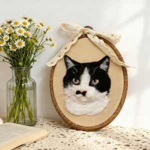 Peut inclure: Un tambour à broder ovale en bois contenant un portrait en laine feutrée d'un chat noir et blanc au nez rose. Le tambour est orné d'un ruban en dentelle couleur crème noué en nœud sur le dessus, encadrant le visage texturé du félin.