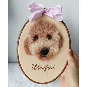 May include: An oval wooden embroidery hoop containing a needle-felted portrait of a curly-haired golden dog. The dog has dark eyes and a black nose. Below the dog, the name Winifred is embroidered in brown thread. A light pink fabric bow is attached.