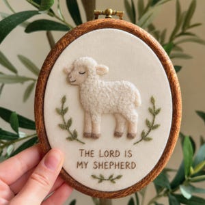 May include: An oval embroidery hoop with a wooden frame containing cream-colored fabric. The fabric features a needle-felted white lamb, two green embroidered leafy branches, and the text "THE LORD IS MY SHEPHERD" stitched in brown thread.