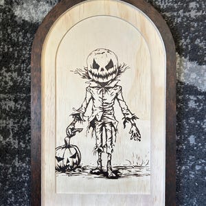 May include: A wooden Halloween-themed wall art piece. The artwork features a black and white illustration of a scarecrow with a carved pumpkin head and a matching pumpkin. The artwork is framed with a dark brown wooden frame.