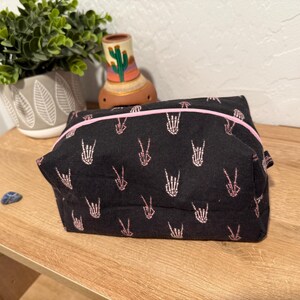May include: Black zippered pouch with a pink zipper and a repeating pattern of pink skeleton hands making the peace sign.