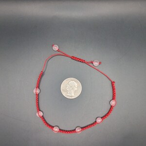 May include: A red macrame bracelet with six pink quartz beads. The bracelet is adjustable and features a sliding knot closure. A quarter is next to the bracelet for size reference.