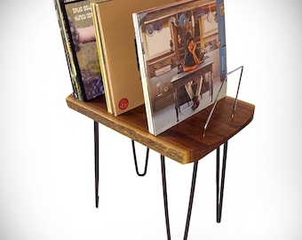 May include: A wooden record stand with black hairpin legs. The stand has a shelf for storing records and a metal rack for holding records upright. The stand is made of natural wood and has a rustic look.
