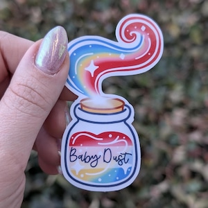 May include: A sticker of a glass jar with a cork lid filled with colorful glitter and a rainbow-colored swirl of smoke coming out of the top. The jar has the text "Baby Dust" written on it.