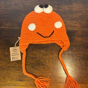 Könnte beinhalten: Orangefarbene Häkelmütze mit einem lächelnden Gesicht, zwei weißen Augen mit schwarzen Pupillen und zwei langen orangenen Bommeln. Die Mütze ist handgefertigt und hat ein Etikett mit der Aufschrift "Schlafenszeit Kunsthandwerk".