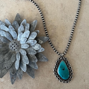 May include: A necklace featuring a teardrop-shaped turquoise stone set in a silver frame with a scalloped edge. The pendant hangs from a silver bead chain that transitions into a black leather cord. A metal flower ornament is positioned nearby.