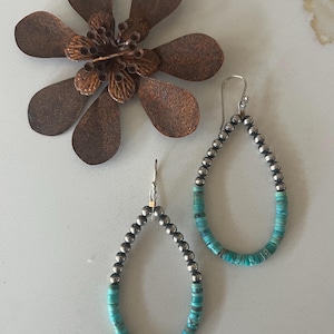 May include: A pair of teardrop-shaped earrings featuring a combination of polished silver-tone beads and small, flat turquoise-colored discs. The earrings hang from silver-tone hooks. The text @DogAndDragonfly is visible at the bottom.