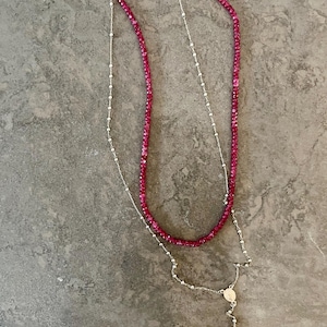 Puede incluir: Dos collares sobre una superficie gris. Un collar es una hebra de pequeñas cuentas de color rosa brillante. El otro es una cadena de plata con un colgante de cruz y un pequeño colgante circular. Los collares están superpuestos.