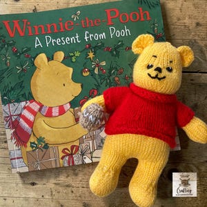 May include: A yellow knitted bear toy wearing a red sweater and holding a small brown honey pot, positioned next to a Winnie-the-Pooh: A Present from Pooh book. The bear has embroidered facial features and the honey pot has the word HUNNY on it.