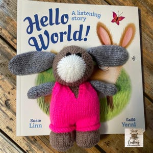 May include: A hand-knitted brown and gray plush toy with a white muzzle, wearing bright pink overalls, positioned on top of a children's book titled Hello World! A listening story by Susie Linn, featuring an illustration of a rabbit and a butterfly.