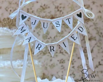 May include: A cake topper featuring two rows of white triangular fabric pennants suspended between two wooden sticks. The pennants display the black text JUST MARRIED with black heart icons at the ends. White satin ribbons are tied to the top.