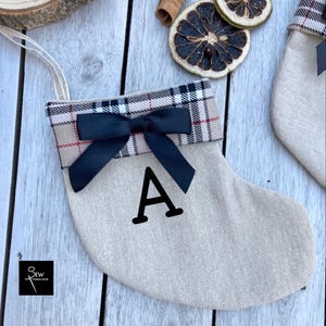 May include: A beige fabric Christmas stocking featuring a cuff with a tan, black, and red plaid pattern. A black satin ribbon bow is centred on the cuff. The letter A is printed in black on the stocking body. A small logo reads Sew Fabulous.