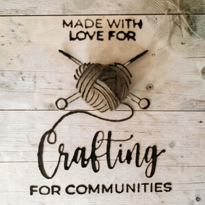 May include: A light-colored wooden sign featuring the burned-in text MADE WITH LOVE FOR, followed by a central graphic of a yarn ball shaped like a heart with two crossed knitting needles, and the text Crafting FOR COMMUNITIES below it.