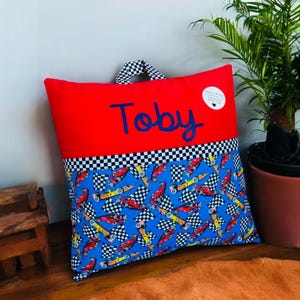 May include: Red and blue personalized pillow with the name Toby embroidered on the front. The back of the pillow features a checkered pattern and a blue fabric with a repeated pattern of race cars and checkered flags.