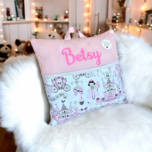 May include: A pink and white personalized pillow with a princess theme. The pillow has a pocket on the back and the name "Betsy" is embroidered on the front.