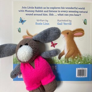 May include: A knitted brown rabbit toy with long gray ears and a white muzzle, wearing a bright neon pink vest. It is placed over a children's book titled Join Little Rabbit as he explores his wonderful world with Mummy Rabbit and listens to every amazing natural sound around him. Shh ... what can you hear? Written by Susie Linn, Illustrated by Gail Yerrill.