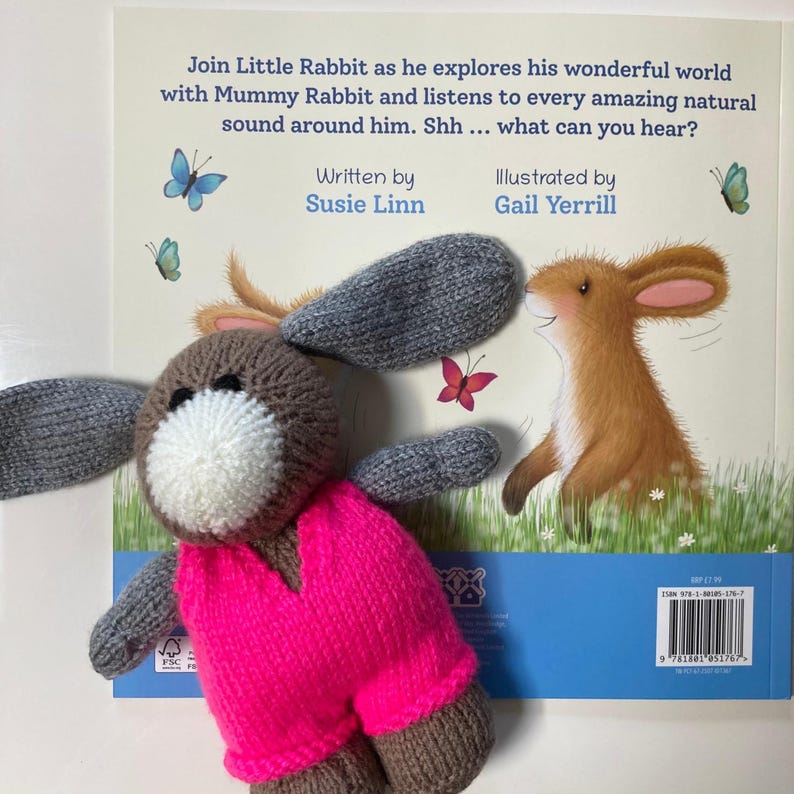 May include: A knitted brown rabbit toy with long gray ears and a white muzzle, wearing a bright neon pink vest. It is placed over a children's book titled Join Little Rabbit as he explores his wonderful world with Mummy Rabbit and listens to every amazing natural sound around him. Shh ... what can you hear? Written by Susie Linn, Illustrated by Gail Yerrill.
