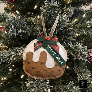 May include: A felt Christmas pudding ornament featuring a brown base with white icing, decorated with a red and green plaid ribbon, green holly leaves, and three red berries. A green tag attached to the ribbon displays the text Merry Xmas.