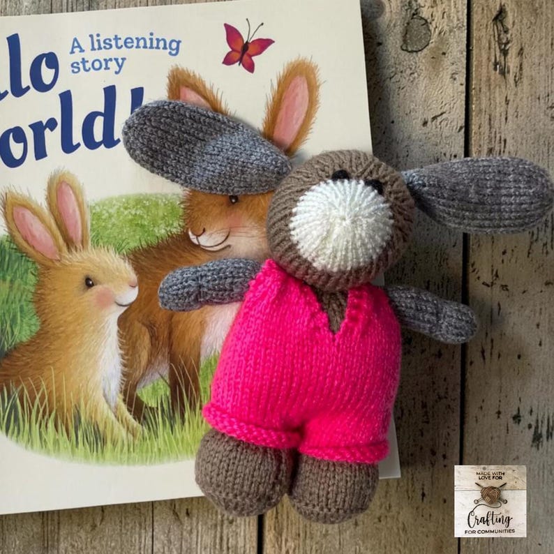 May include: A hand-knitted stuffed animal shaped like a rabbit with long gray ears, a brown head, a white muzzle, and a bright pink jumpsuit. The rabbit is positioned in front of a book titled Hello World A listening story with a rabbit illustration.