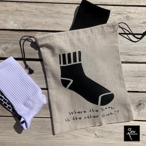 May include: A beige drawstring bag made of woven fabric featuring a black graphic of a sock with striped detailing and the printed text "Where the F*** is the other Sock?!" accompanied by a white sock and a black sock partially inside the bag.