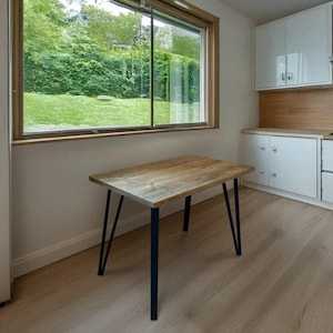 Könnte beinhalten: Ein rechteckiger Holztisch mit natürlicher Oberfläche und schwarzen Metall-Haarnadelbeinen. Der Tisch steht in einem Raum mit einem großen Fenster mit Blick auf einen grünen Garten. Der Tisch ist etwa 122 cm lang.