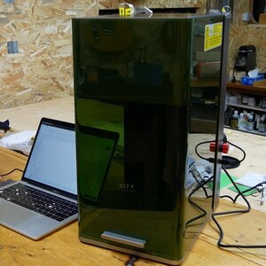 May include: A dark green rectangular machine with the text "ZO-1" on the front. A laptop and a tablet are visible to the left of the machine. The machine has a silver handle at the bottom and a black power cord.
