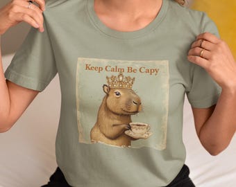 May include: A light green t-shirt with a graphic of a capybara wearing a crown and holding a teacup. The text "Keep Calm Be Capy" is printed on the shirt.