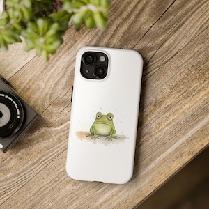 May include: White phone case with a green frog illustration. The frog has a grumpy expression and is sitting on a brown surface with watercolor splatters.