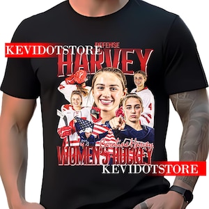 以下が含まれることがあります： 女性のグラフィックと赤い文字で「HARVEY」と書かれた黒いTシャツ。上部に「DEFENSE」、下部に「WOMEN'S HOCKEY」と書かれています。シャツには赤い文字で「KEVIDOTSTORE」も含まれています。