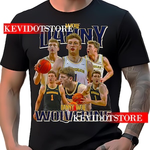 Puede incluir: Camiseta negra con un gráfico de jugadores de baloncesto que llevan camisetas de la Universidad de Michigan. El texto "Danny Wolf" y "Wolverine" está impreso en la camiseta.