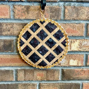 May include: A decorative pie-shaped wall hanging with a brown and black fabric design. The pie crust is a gold color with a scalloped edge.