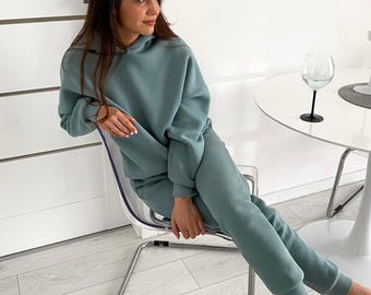 May include: A woman wearing a light blue hooded sweatshirt and matching sweatpants. She is sitting on a clear plastic chair with her legs crossed. The chair is on a light wood floor.