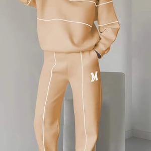 May include: A two-piece outfit in a light brown color with white trim. The top has a mock neck and a zipper closure. The pants have a straight leg and a drawstring waist. Both pieces feature the letter "M" embroidered on the front.