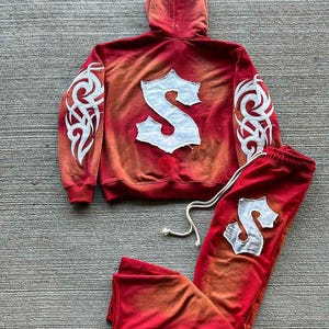 May include: Red and orange hooded sweatshirt and sweatpants set with a white tribal design on the sleeves and a large white letter "S" on the back of the sweatshirt and one leg of the sweatpants.