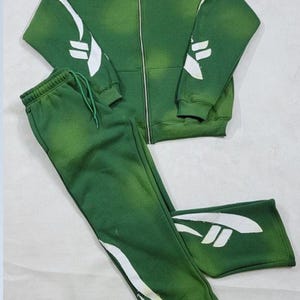 May include: A green hooded sweatshirt and matching sweatpants with white abstract designs. The sweatshirt has a zipper closure.