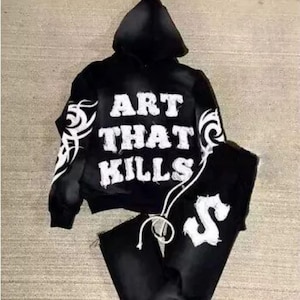 Pode incluir: Camisola de moletom com capuz preta e calças de moletom combinando com a frase "ART THAT KILLS" e um design tribal impresso em branco.