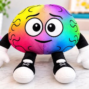 May include: A plush brain toy featuring a vibrant rainbow gradient surface with black embroidered brain fold patterns. The character has large white eyes with black pupils, a simple black smile, black arms with white gloves, and black sneakers.