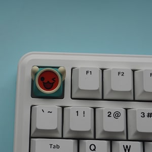 May include: A white mechanical keyboard with a custom keycap featuring a red and teal cartoon face. A black ESC key is separate. The keyboard has white keys with black lettering. The background is a light blue.