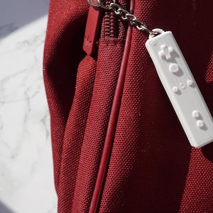 Puede incluir: Un llavero de plástico blanco con forma de mando de videojuego. El llavero está unido a una bolsa roja con un logotipo blanco.