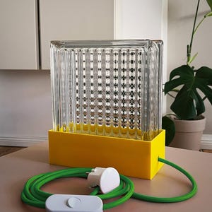 K&ouml;nnte beinhalten: Eine rechteckige Tischlampe aus gelbem und klarem Glas mit einem gr&uuml;nen Kabel und einem wei&szlig;en Schalter. Die Lampe hat einen rechteckigen Sockel und einen klaren Glasschirm mit einem strukturierten Muster.