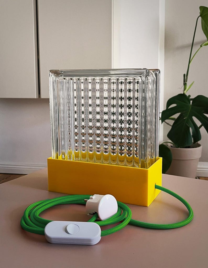 K&ouml;nnte beinhalten: Eine rechteckige Tischlampe aus gelbem und klarem Glas mit einem gr&uuml;nen Kabel und einem wei&szlig;en Schalter. Die Lampe hat einen rechteckigen Sockel und einen klaren Glasschirm mit einem strukturierten Muster.